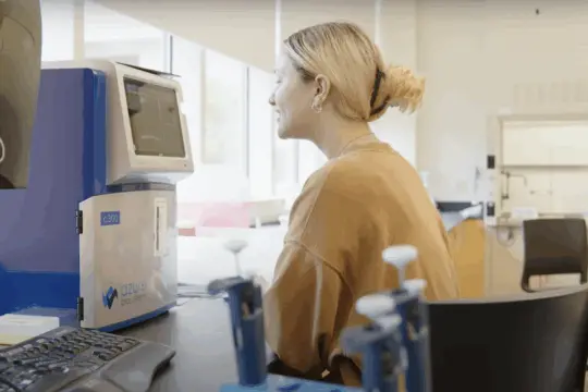 A person with blonde hair is sitting and smiling at a laboratory desk, looking at a device labeled 